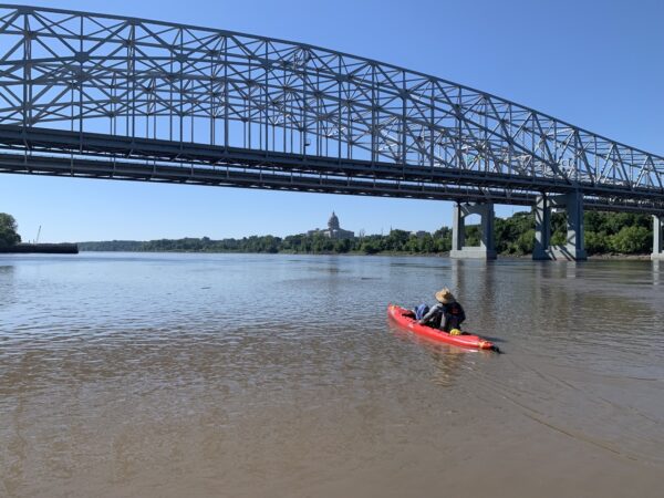 A man in a red kayak on a wide river with a large metal bridge and the MO capitol building in the background.