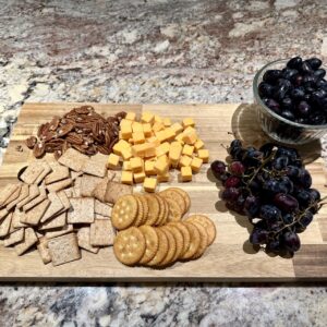 A wooden cutting board with cheese, nuts, crackers, and grapes on a marble countertop.