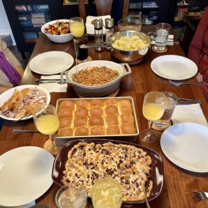 A long wooden dinner table set with a Thanksgiving meal including sweet potato casserole, homemade bread, sliced turkey, green bean casserole, mashed potatoes and gravy, and fruit salad.