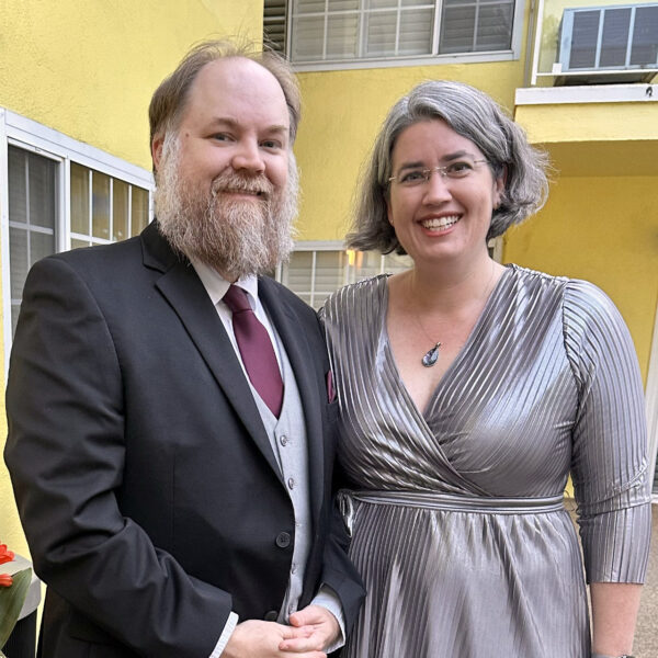 Mr. M in a black suit, silver waistcoat, and maroon tie with me beside him in a shiny silver dress, with yellow walls behind us.
