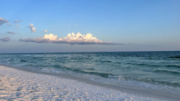 A photo of an empty beach with white sand and small waves and fluffy white clouds in the sky.